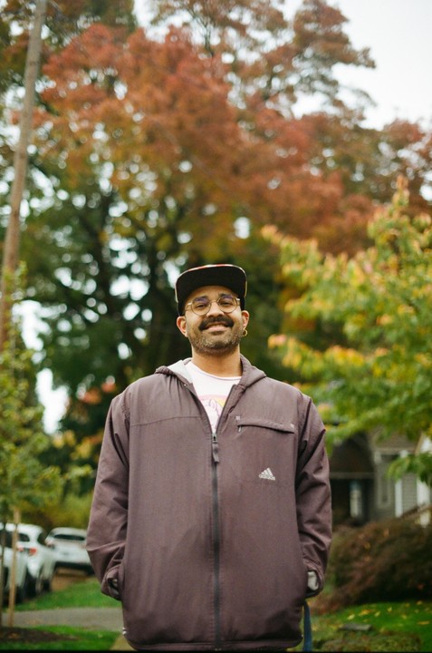 A man wearing a jacket and a cap smiles at the camera with autumn trees in the background.