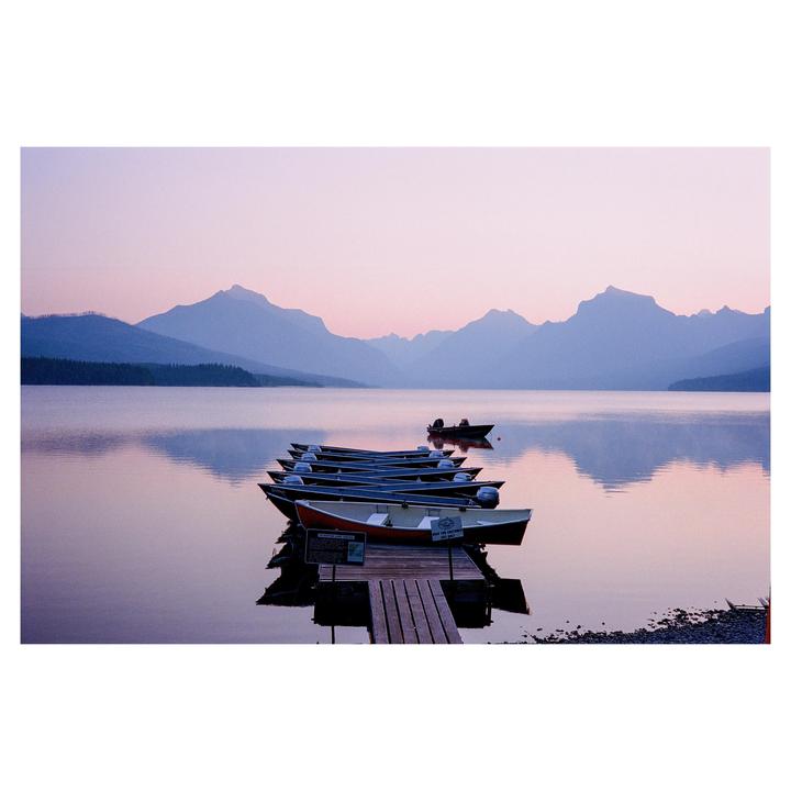 A line of boats is tied to a wooden dock on a lake with mountains in the background at dusk.