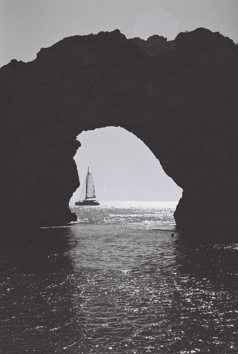 A sailboat passes through a natural rock archway over sparkling water.