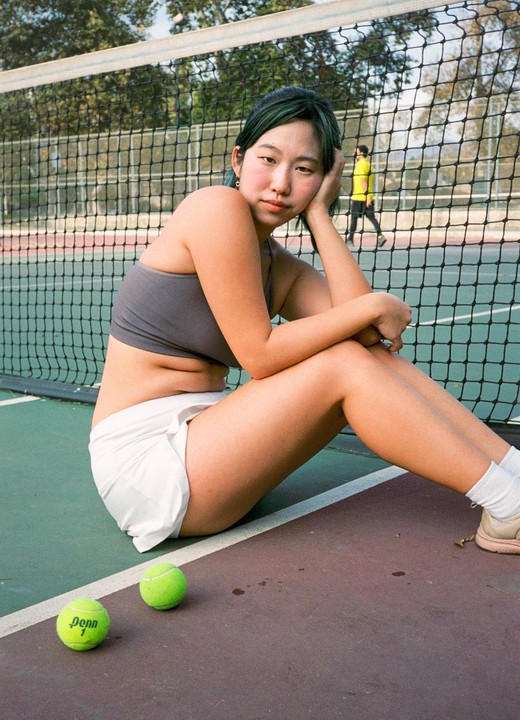 A woman with green hair sits on a tennis court next to two tennis balls.
