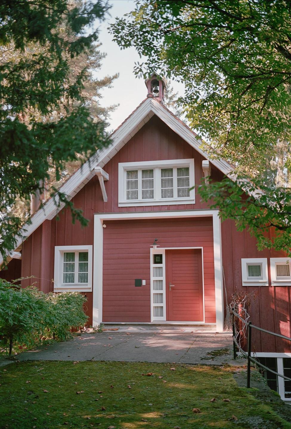 A red wooden house with white trim and a bell on the roof is partially obscured by green foliage.