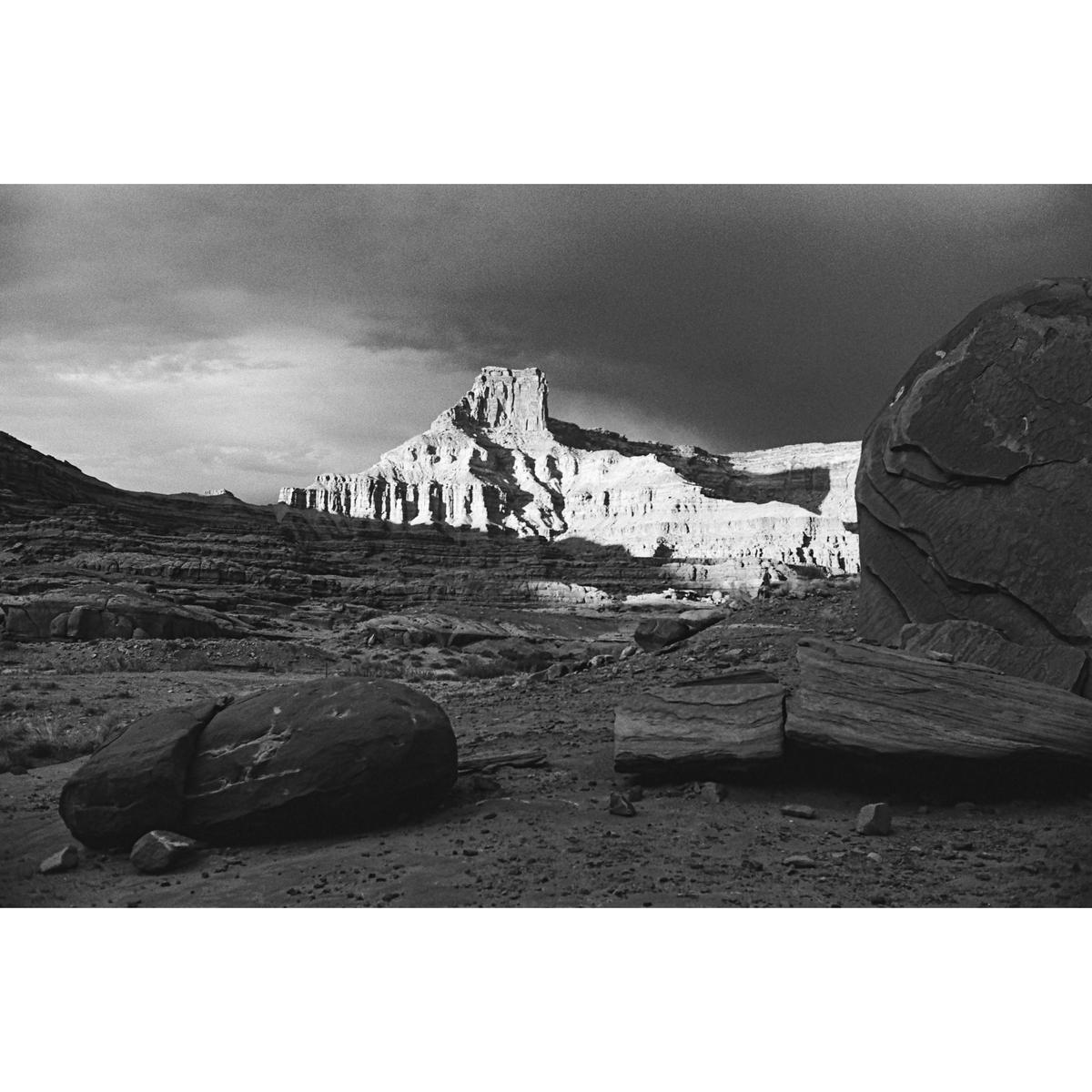 A dramatic black and white landscape shows a sunlit mesa under a dark, cloudy sky, with large boulders in the foreground.