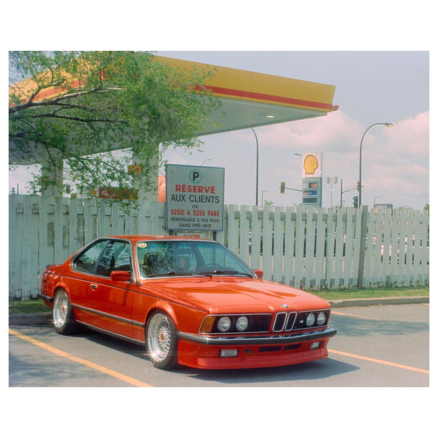 A red car is parked in a lot next to a white fence and a gas station.