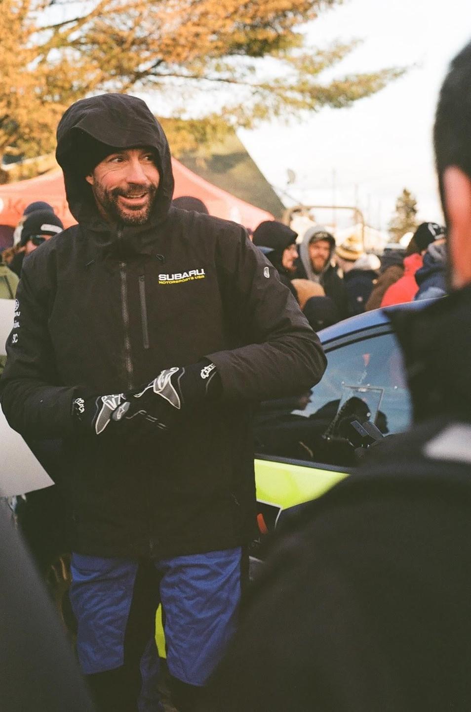 A man in a black jacket and blue pants smiles while standing among a crowd of people.