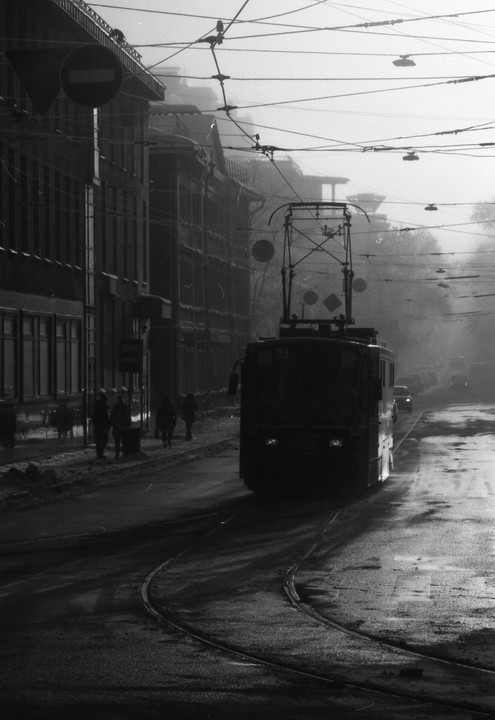 A tram is driving down a street with buildings and people on the sidewalk on a hazy day.