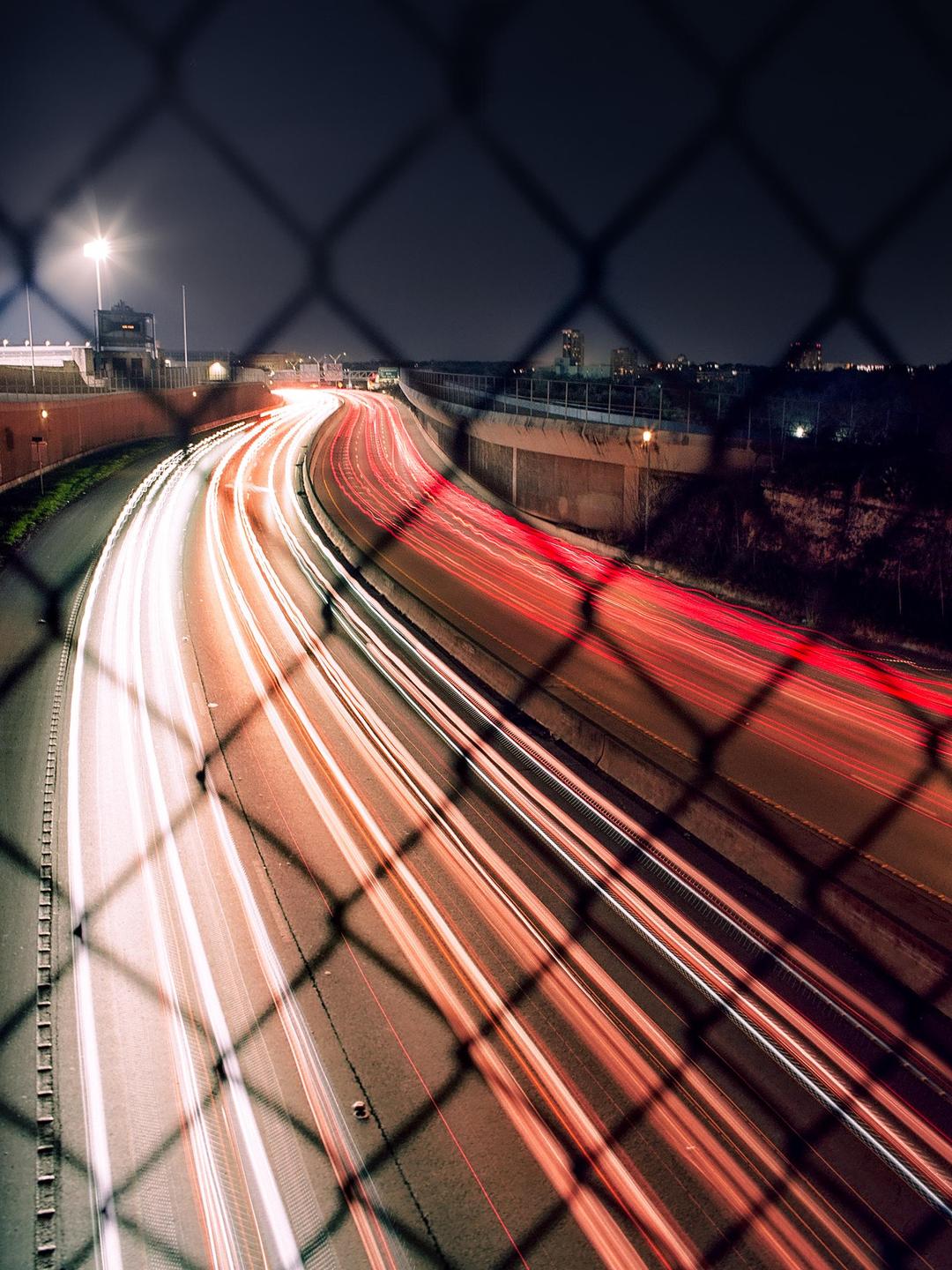 Light trails from vehicles on a highway at night are visible through a chain link fence.