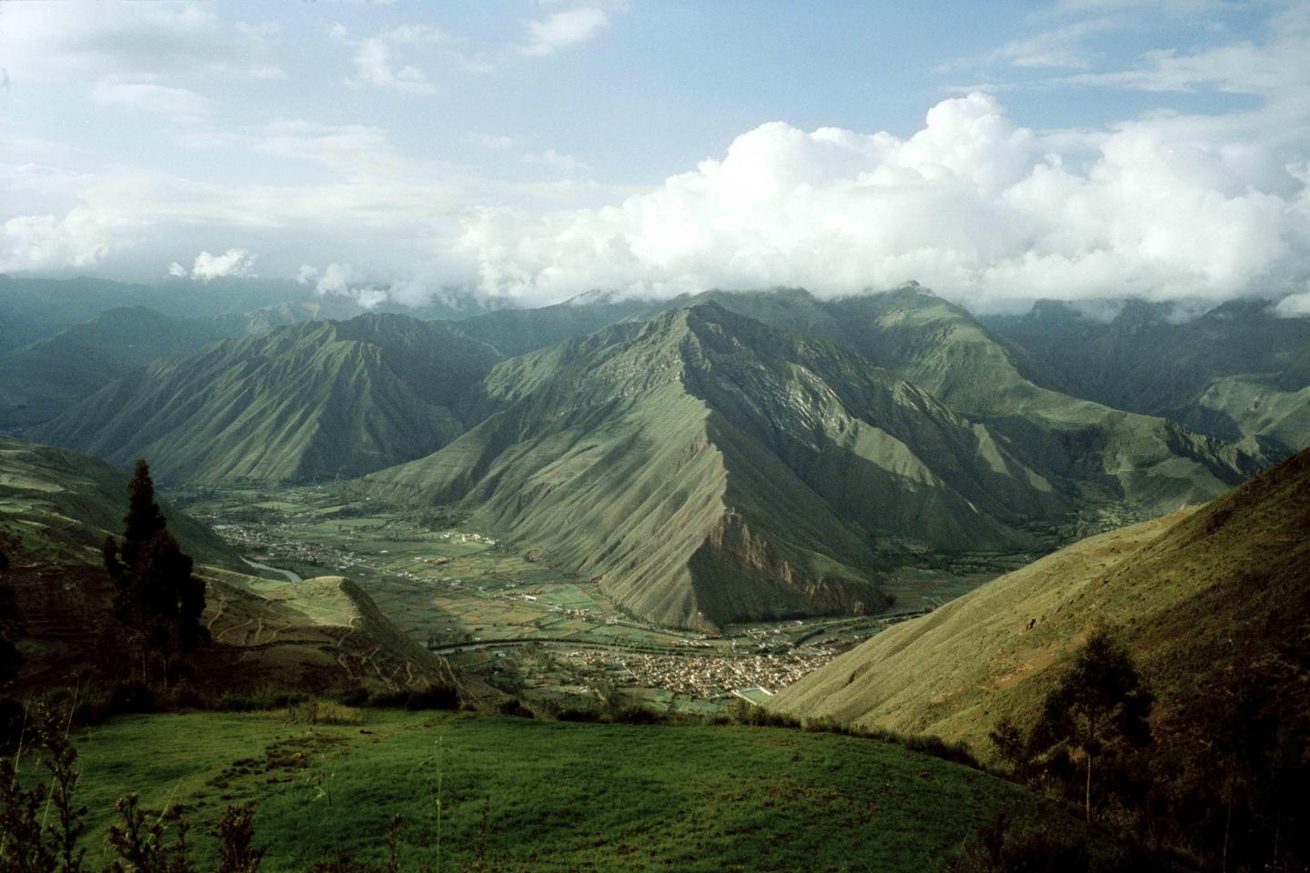 A valley with a town and river is nestled between large mountains under a cloudy sky.
