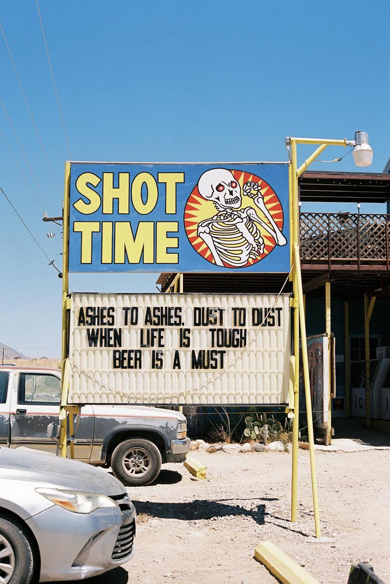 A sign with a skeleton illustration and text about "Shot Time" and "Beer is a Must" stands next to parked vehicles under a clear sky.