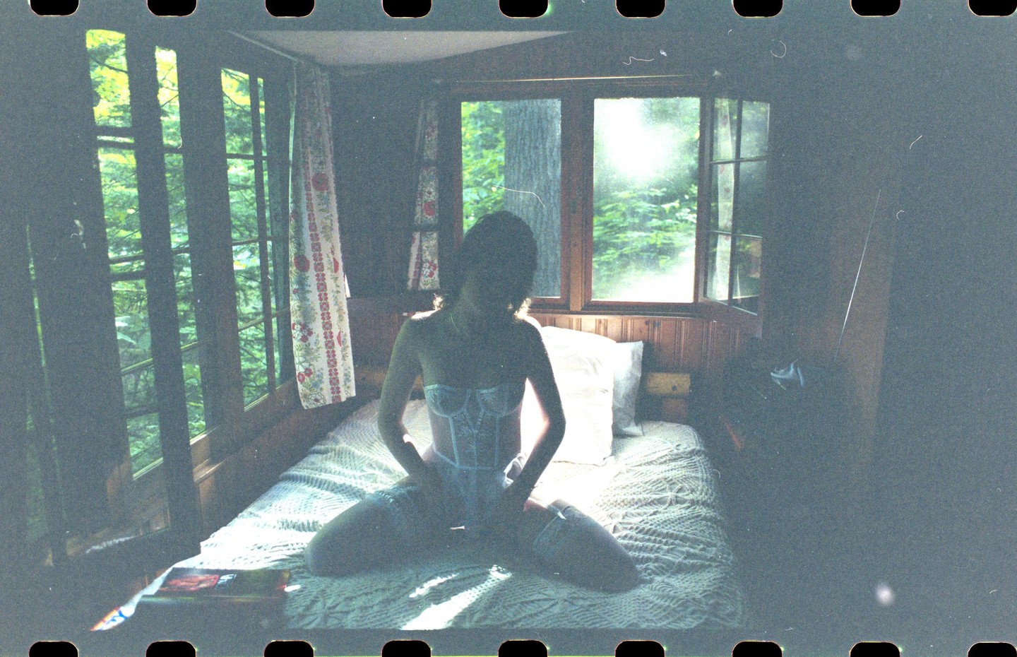 A woman in lingerie kneels on a bed in a cabin with sunlight streaming through the windows.