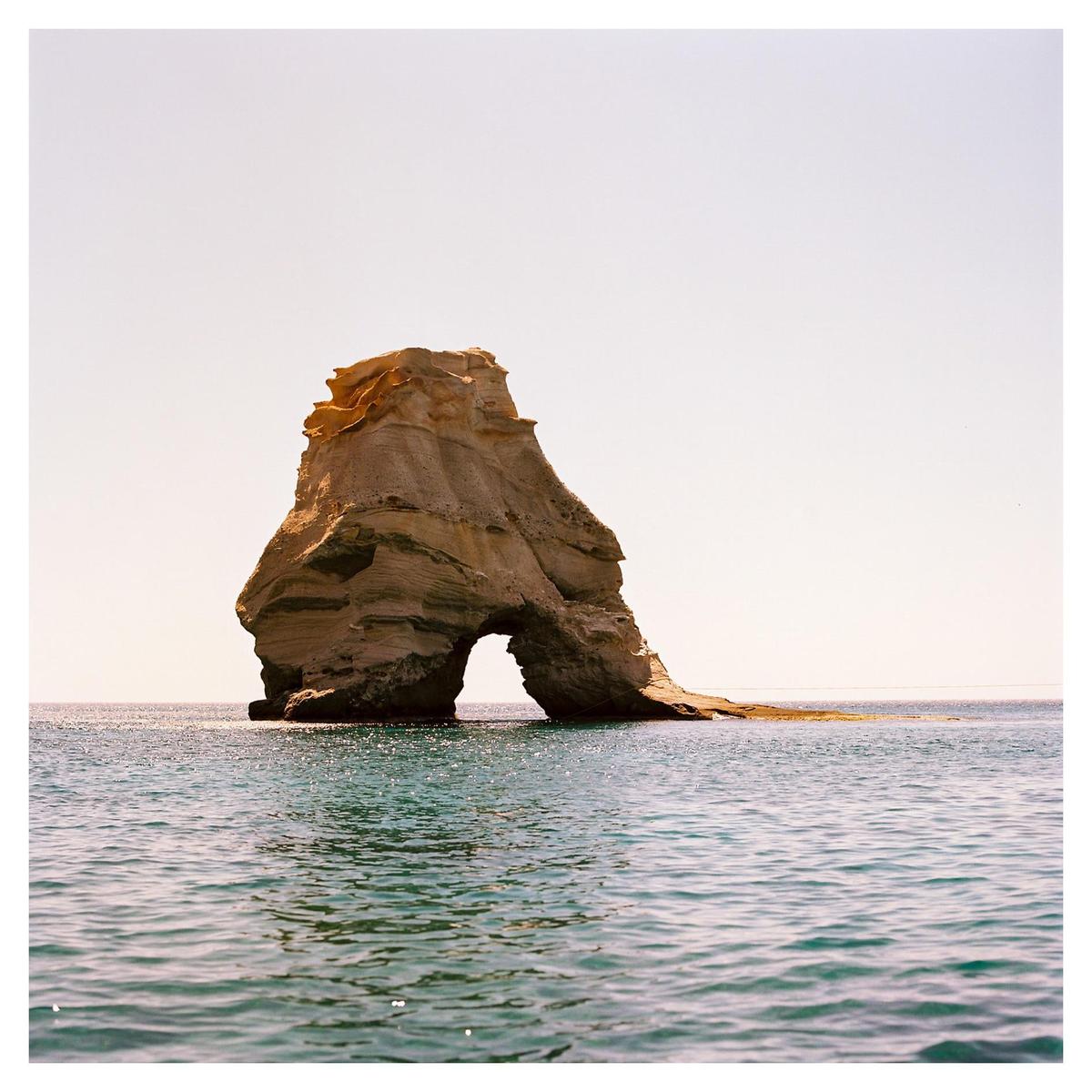 A large rock formation with an archway stands in the middle of the sea under a clear sky.