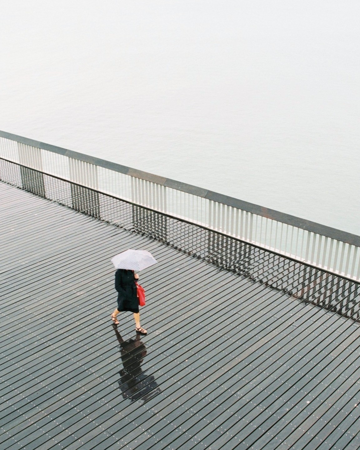 A woman walks with an umbrella on a boardwalk next to water.