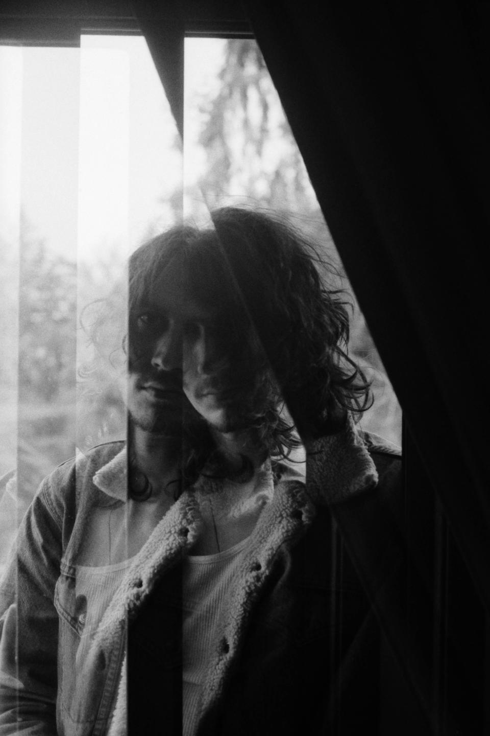 A man with long hair and a beard is visible through a window, with a double exposure effect.