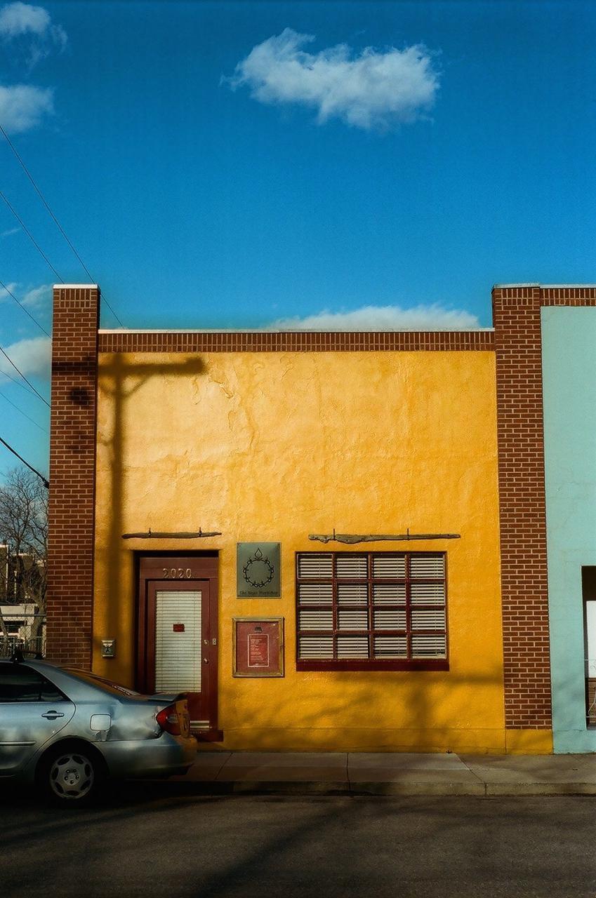 A silver car is parked in front of a yellow building with a red door and a window with blinds, under a bright blue sky with white clouds.