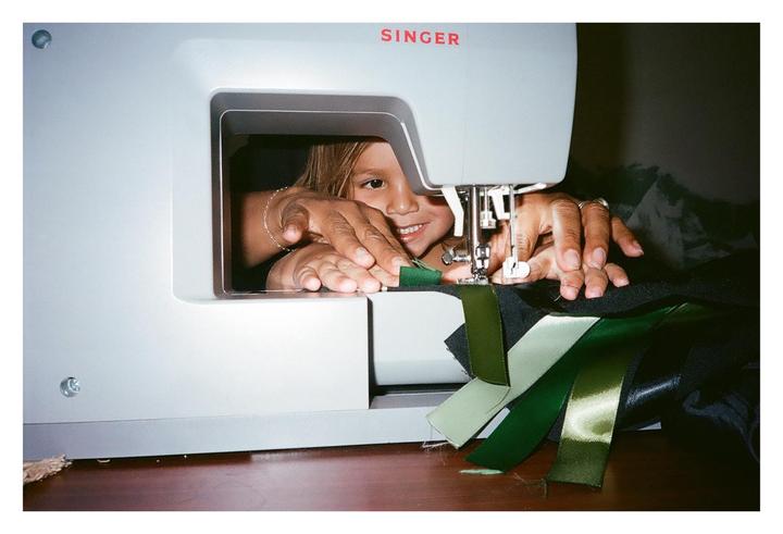 A child smiles while looking through the opening of a sewing machine as an adult's hands guide fabric under the needle.