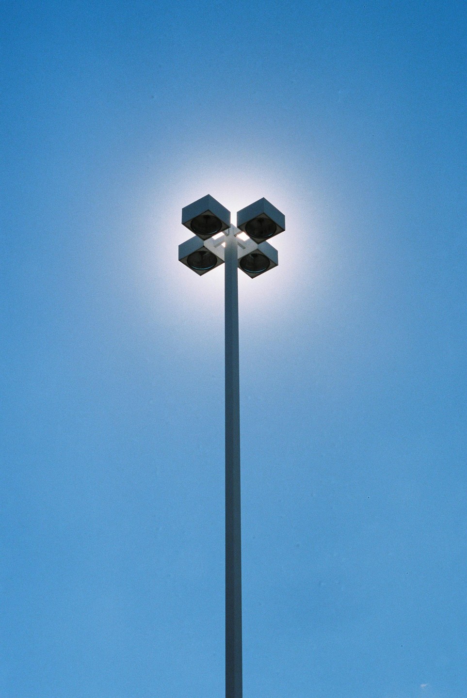 A street light with four square lamps is silhouetted against a bright blue sky.
