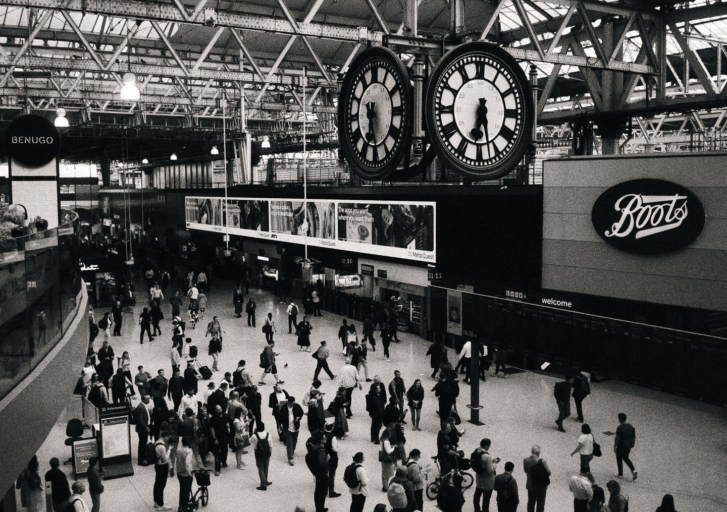 A large crowd of people walk through a train station with two large clocks hanging above them.