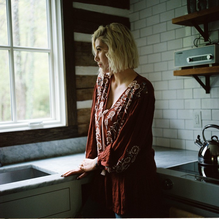 A woman with short blonde hair stands in a kitchen and looks out a window.