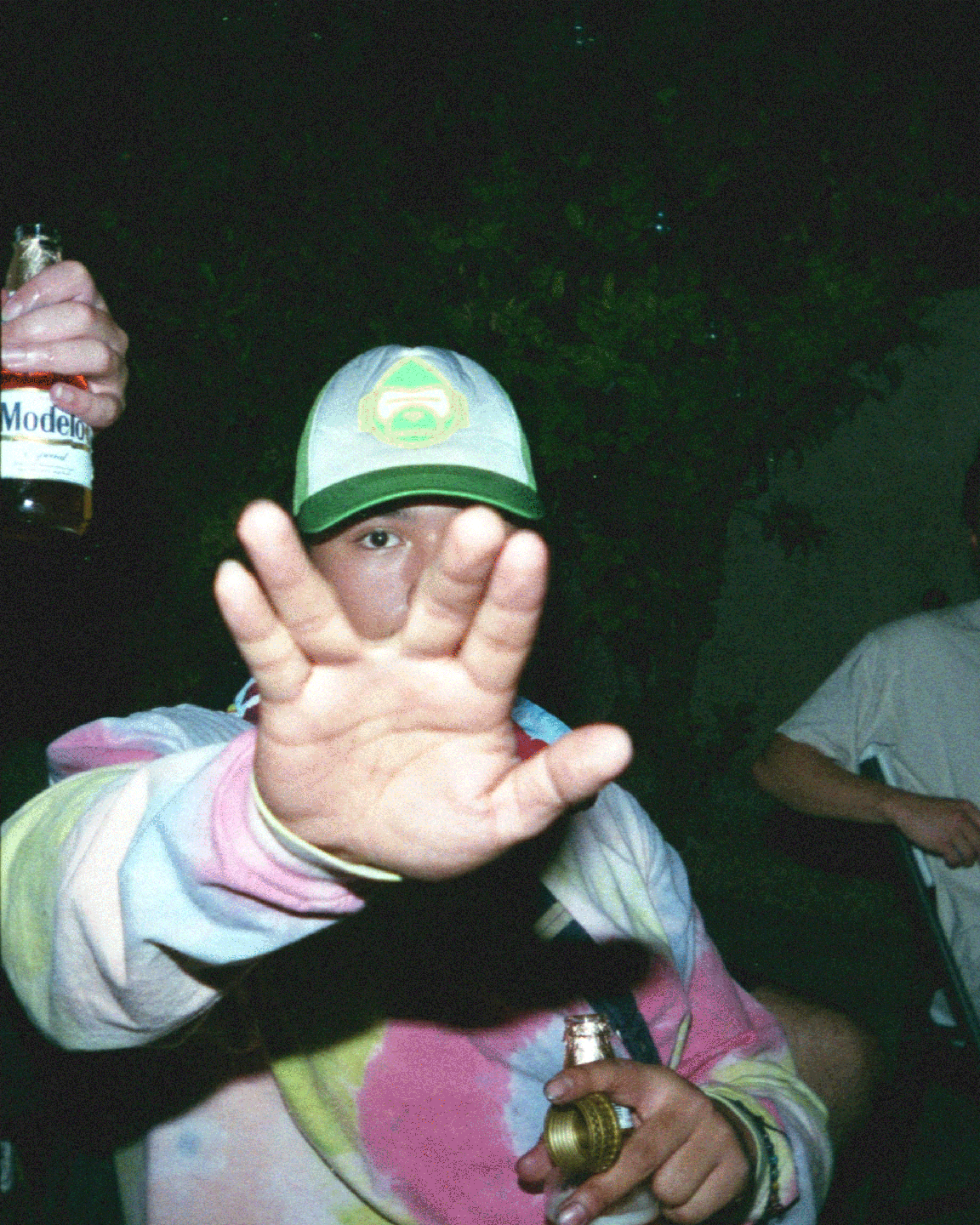 A person wearing a tie-dye hoodie and a cap holds up a hand to the camera while holding a bottle.