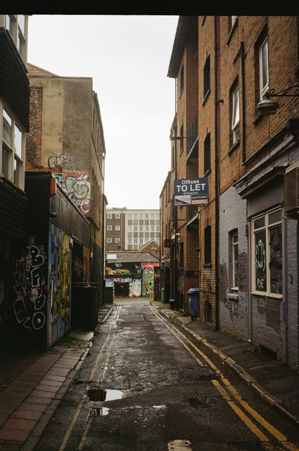 A narrow alleyway with graffiti-covered walls and puddles on the ground leads to a distant building.
