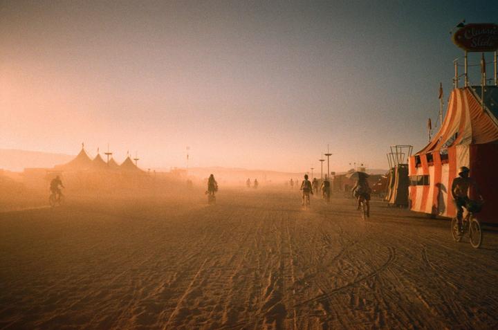 People ride bicycles through a dusty desert landscape with tents and structures under a hazy sky.