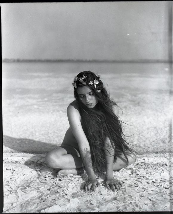 A woman with long hair and a seashell crown kneels on a salt flat.