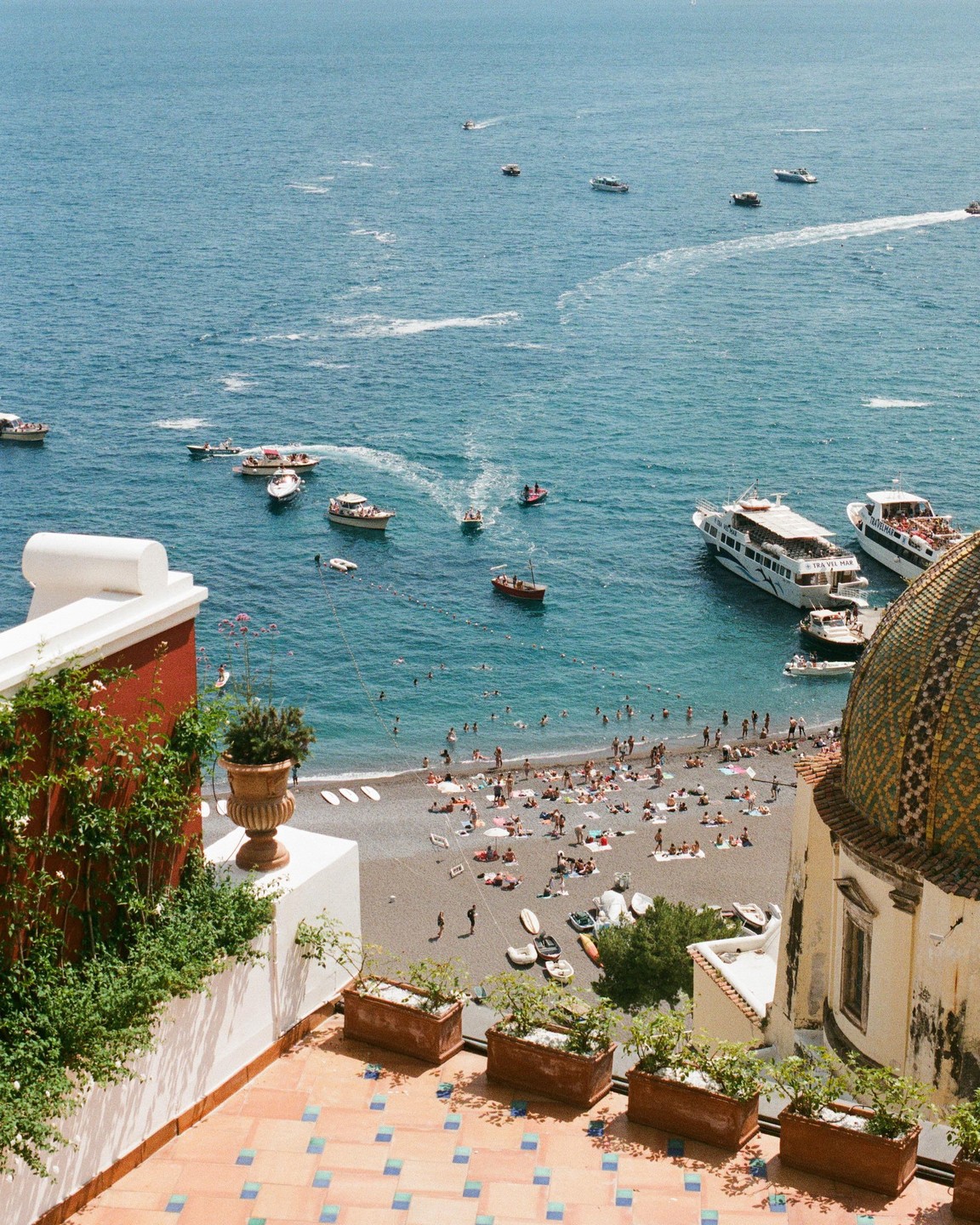 A high-angle view shows a beach crowded with people and boats in the water, with a tiled dome and a terrace in the foreground.