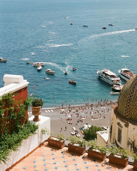 A high-angle view shows a beach crowded with people and boats in the water, with a tiled dome and a terrace in the foreground.