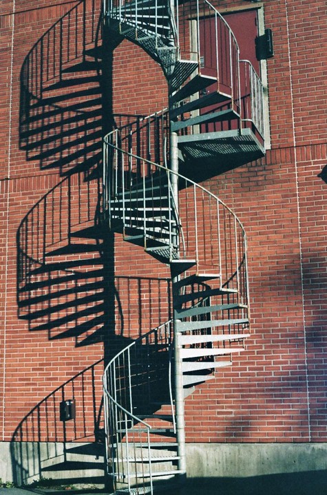 A metal spiral staircase casts a shadow on a red brick wall next to a red door.