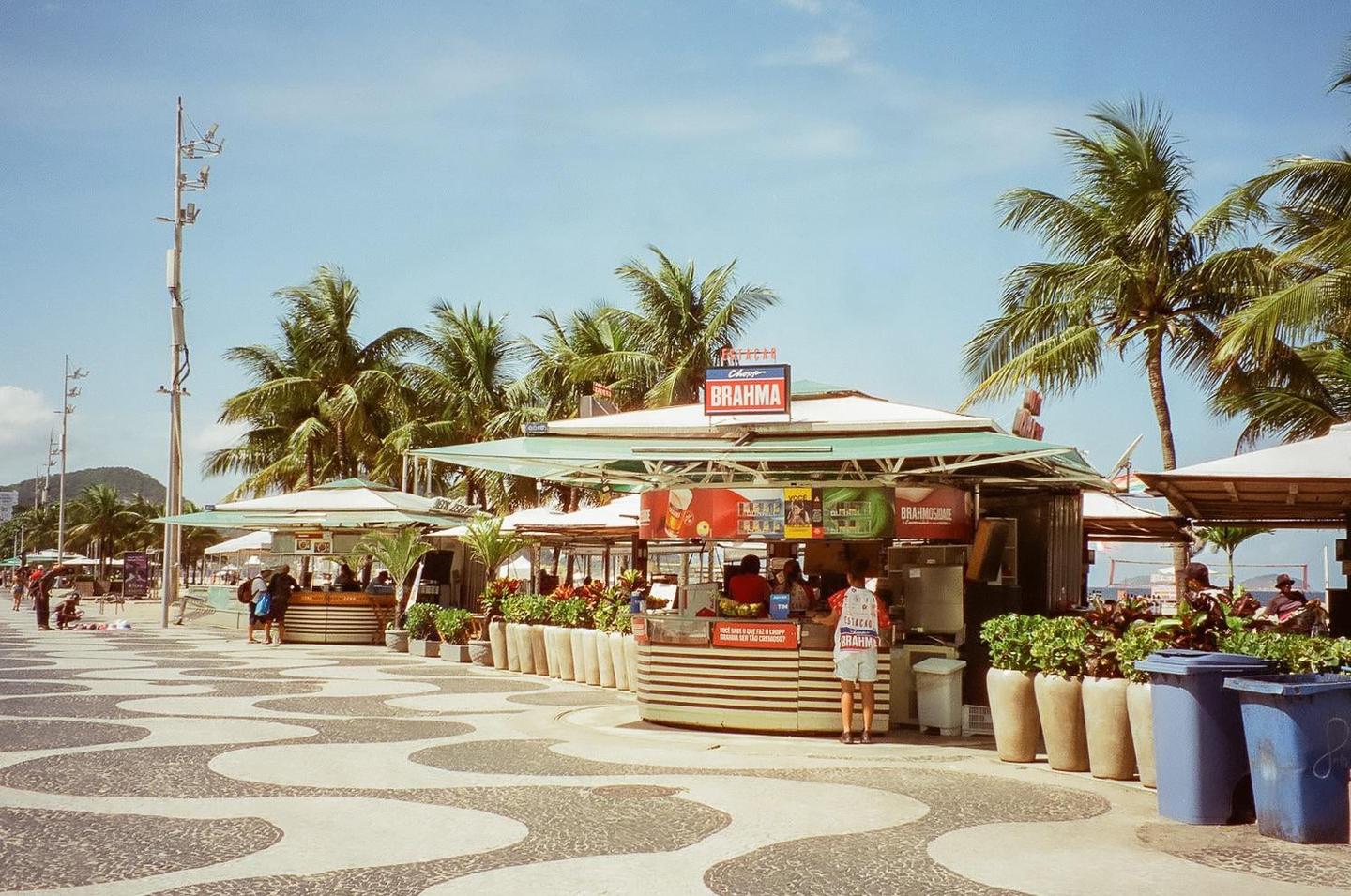 A person stands at a kiosk on a patterned promenade lined with palm trees and other kiosks.