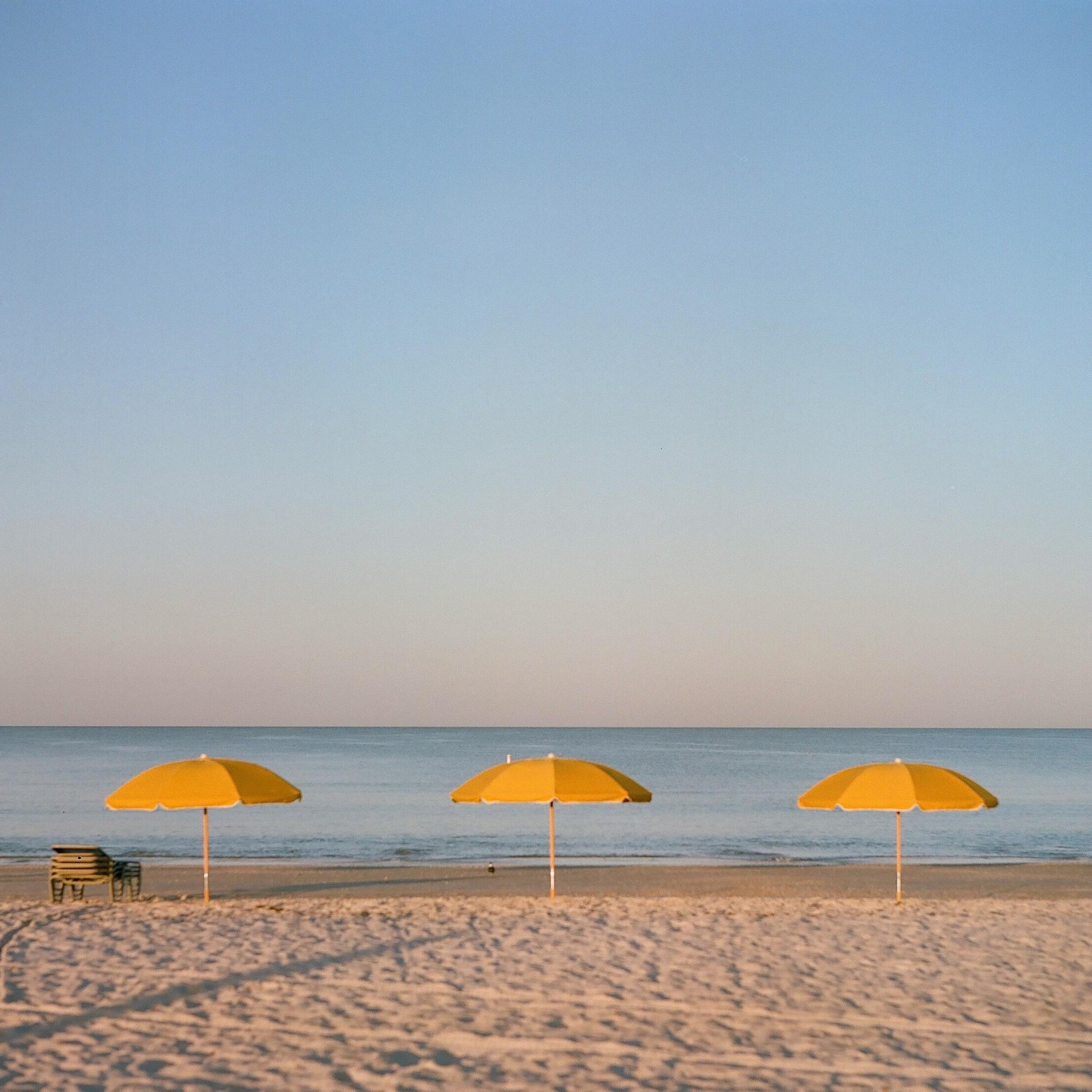 Three yellow beach umbrellas stand on a sandy beach next to the ocean under a clear sky.