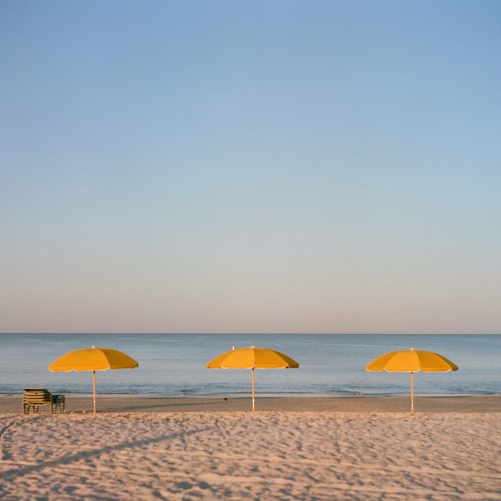 Three yellow beach umbrellas stand on a sandy beach next to the ocean under a clear sky.