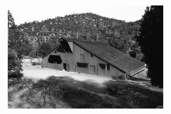 A large wooden barn with a long, sloping roof stands in a rural landscape with a forested hill in the background.