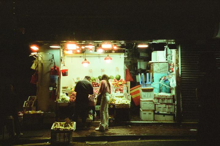 A man and a woman are standing in front of a fruit stall at night.