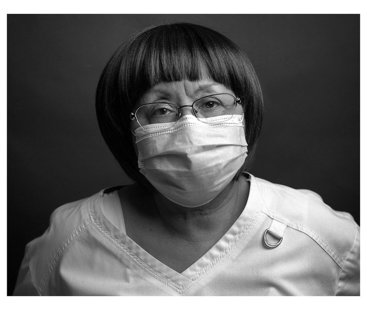 A woman wearing glasses and a surgical mask looks directly at the viewer.