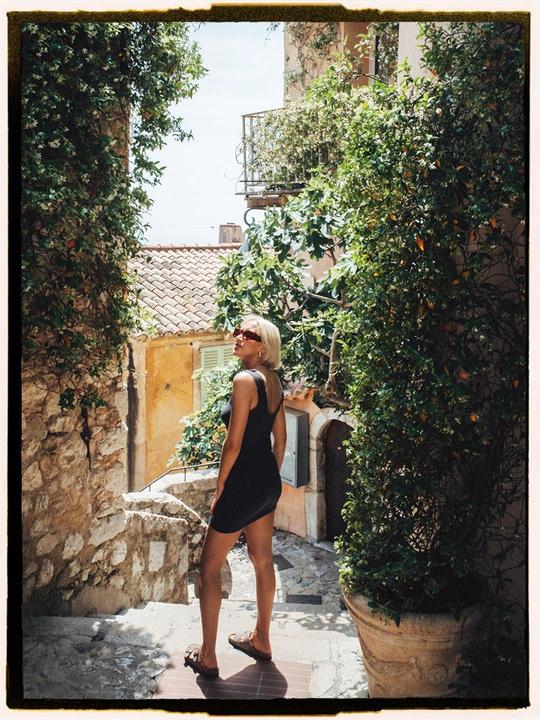 A woman in a black dress stands on stone steps surrounded by lush green foliage and old buildings.