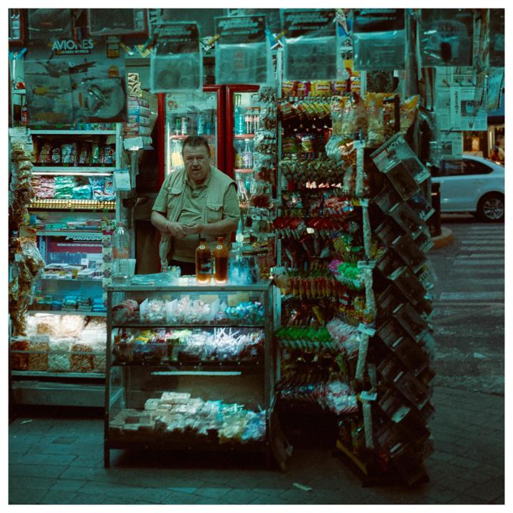 A man stands behind a counter filled with various items at a street stall at night.