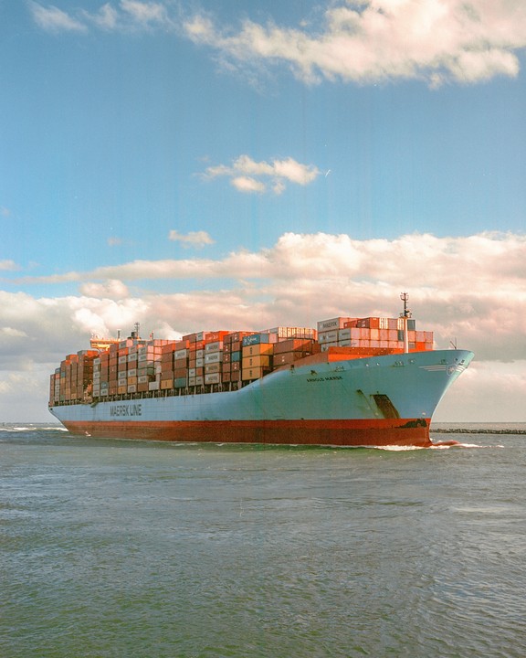 A large cargo ship with many containers on its deck moves through the water under a partly cloudy sky.