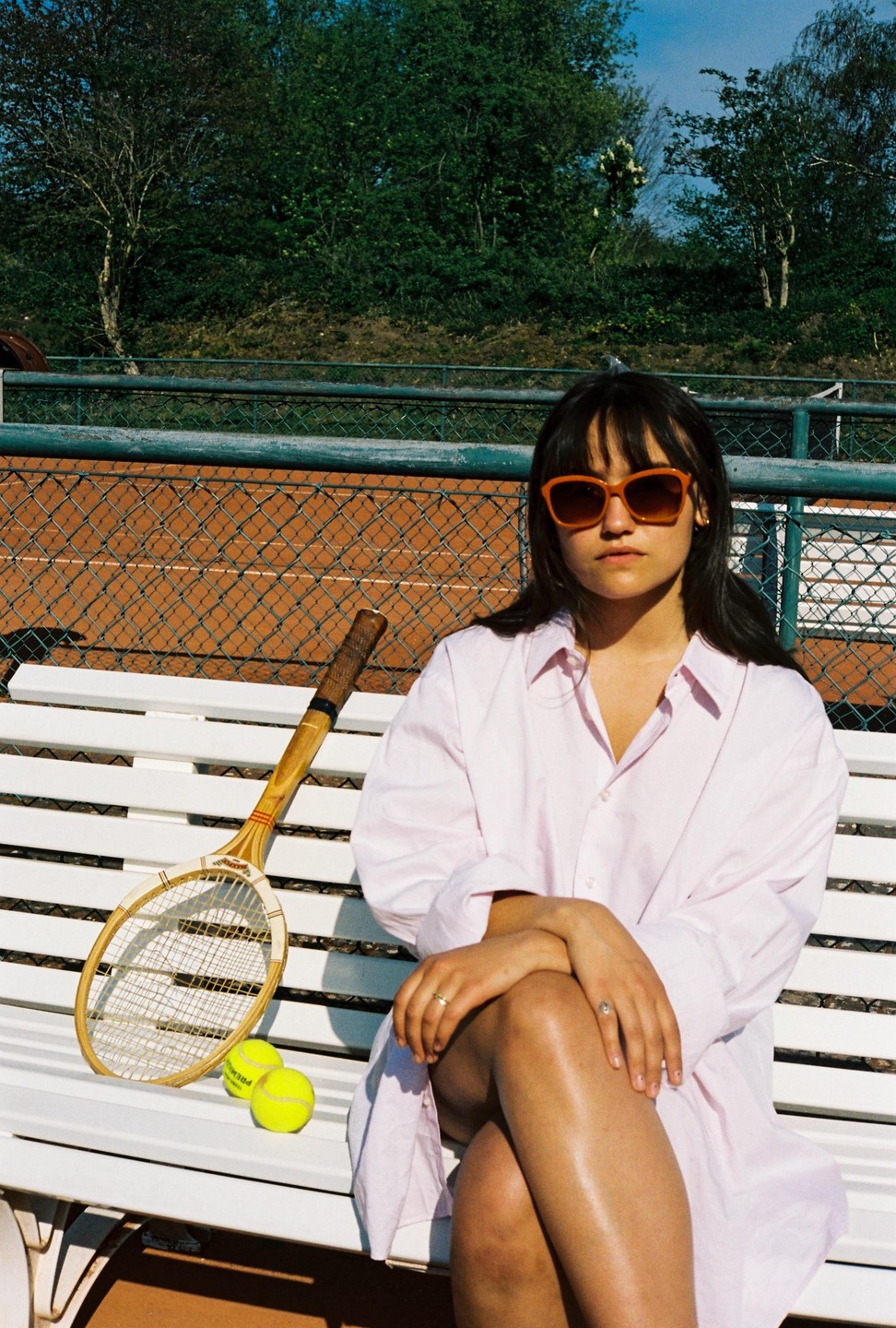A woman wearing sunglasses and a pink shirt sits on a white bench next to a tennis racket and two tennis balls on a sunny day.