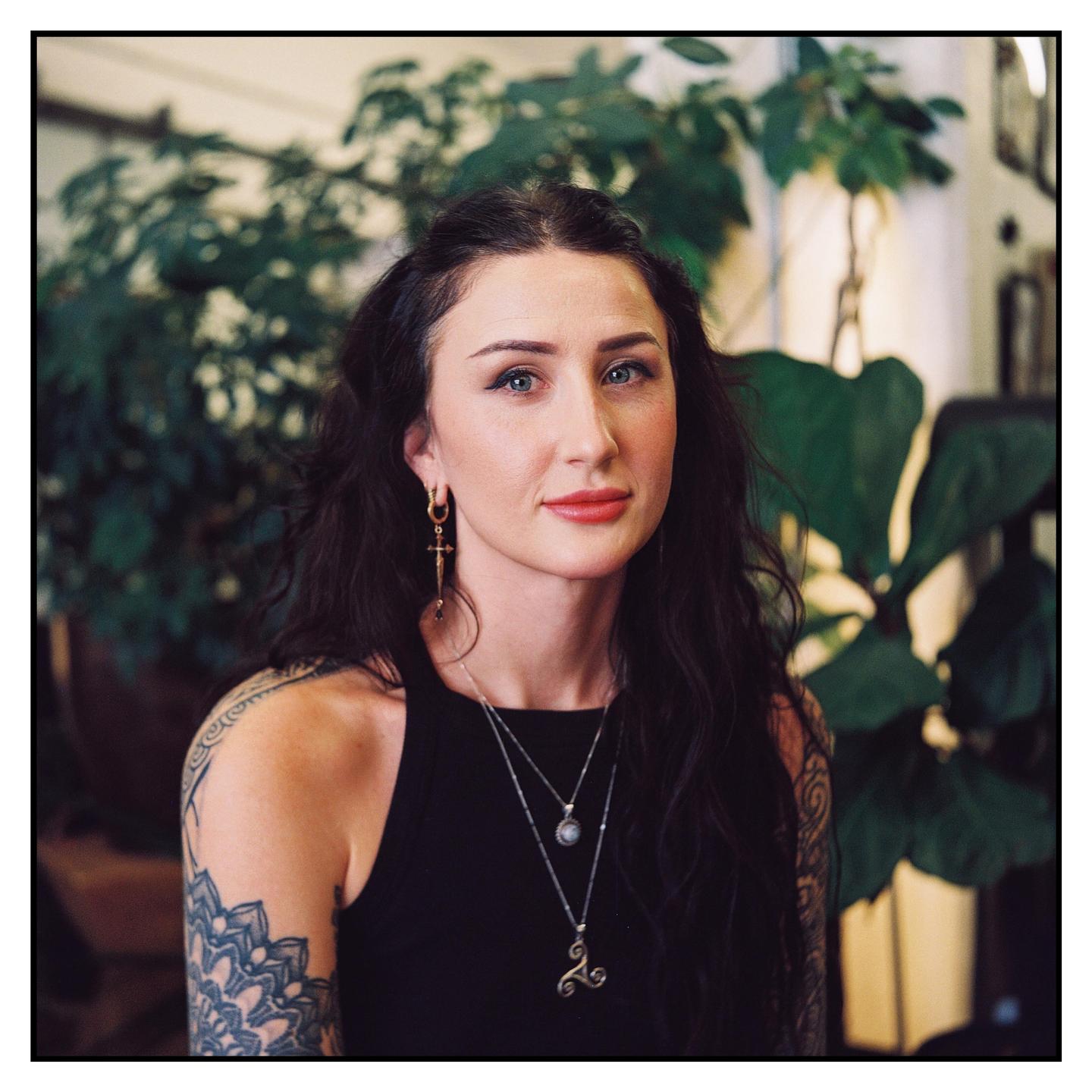 A woman with dark hair and tattoos on her arm looks directly at the viewer.