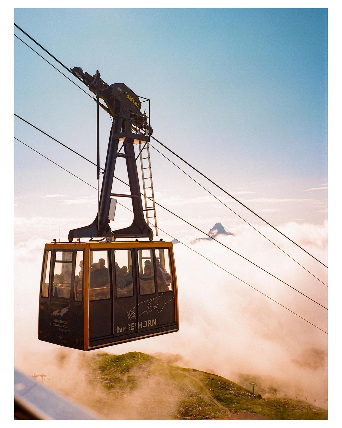 A cable car full of people is suspended over a mountain landscape with clouds and a distant peak.