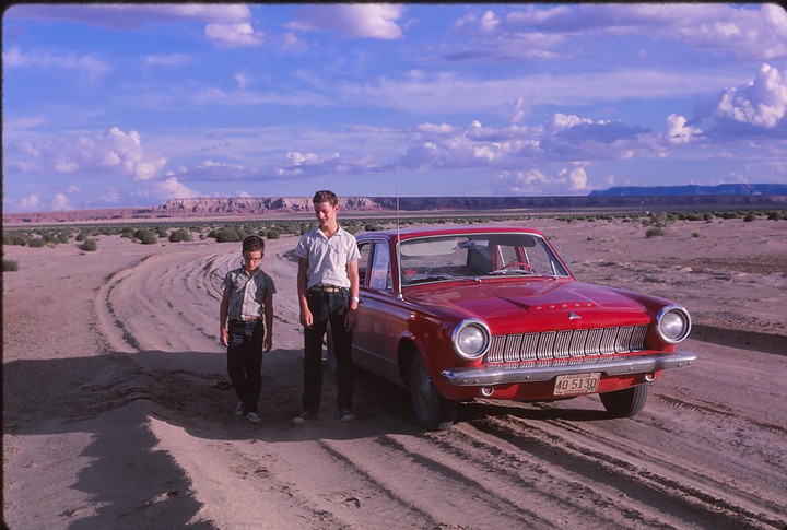 Two boys stand next to a red car on a dirt road in a desert landscape with mountains in the distance.