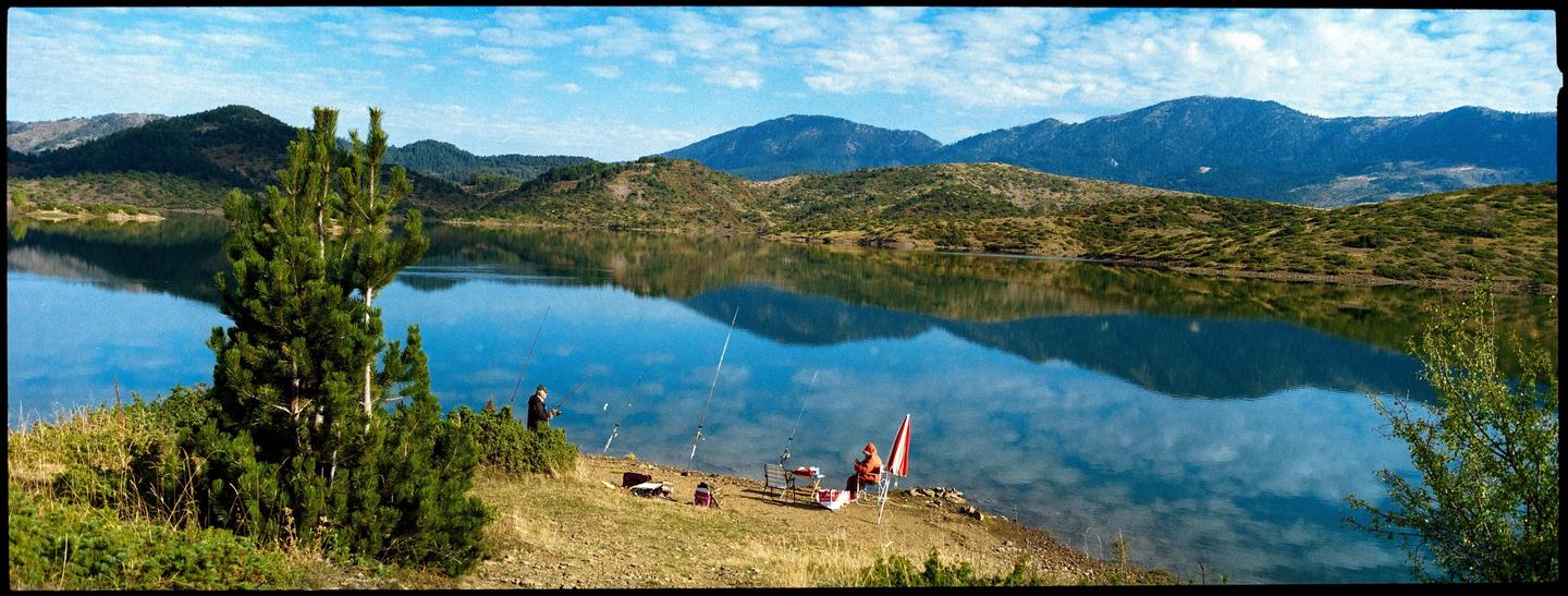 Two men fish on the shore of a lake with mountains in the background.