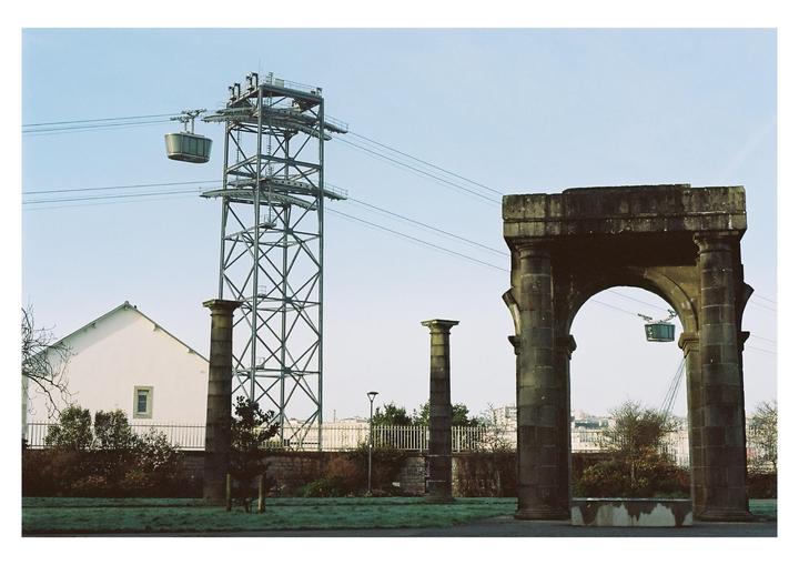 A cable car passes over a park with an archway and a tall metal tower.
