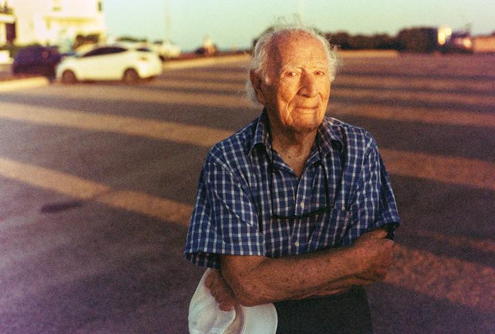 An elderly man in a plaid shirt stands with his arms crossed, holding a white hat, with cars and a street in the background.