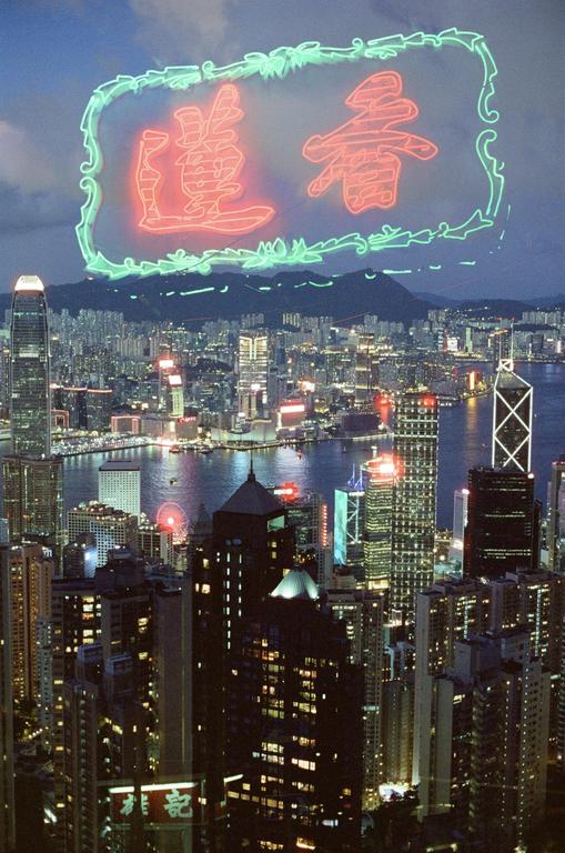 A neon sign with Chinese characters is superimposed over a night cityscape with a body of water and mountains in the distance.
