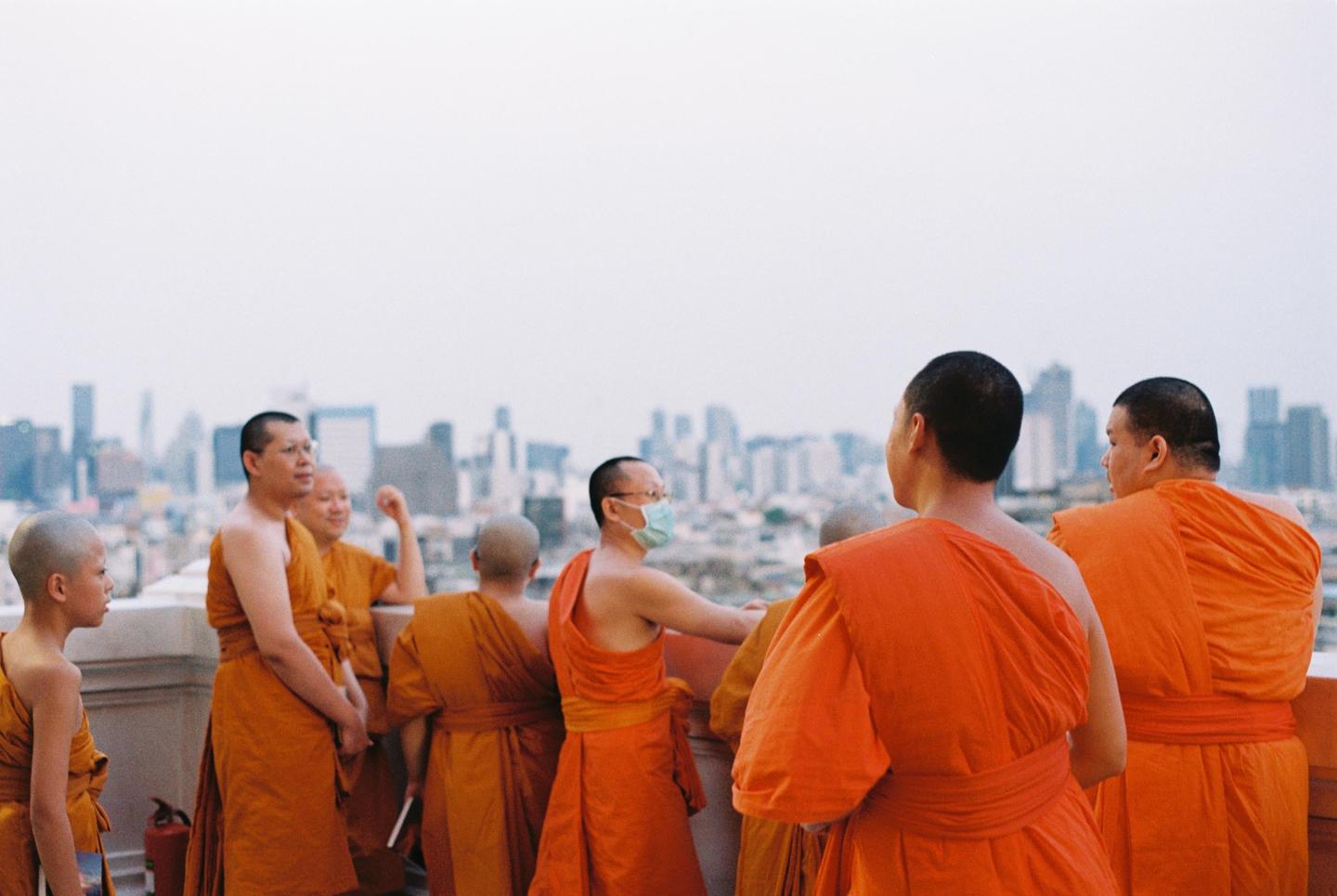 A group of monks in orange robes stands on a rooftop overlooking a city skyline.