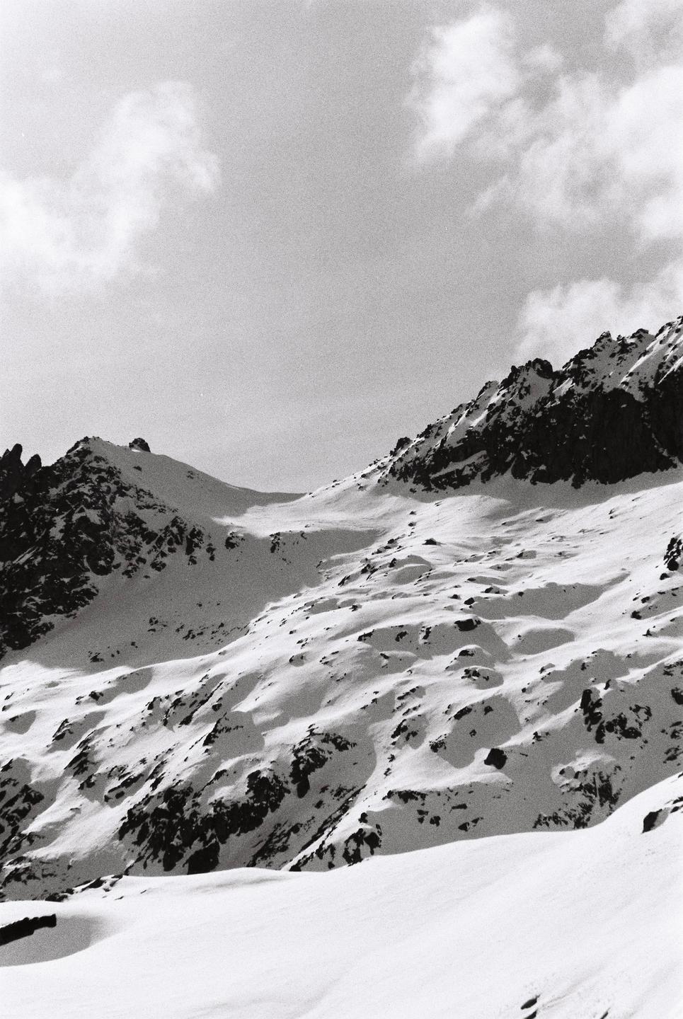 A black and white photograph captures a snow-covered mountain range under a cloudy sky.