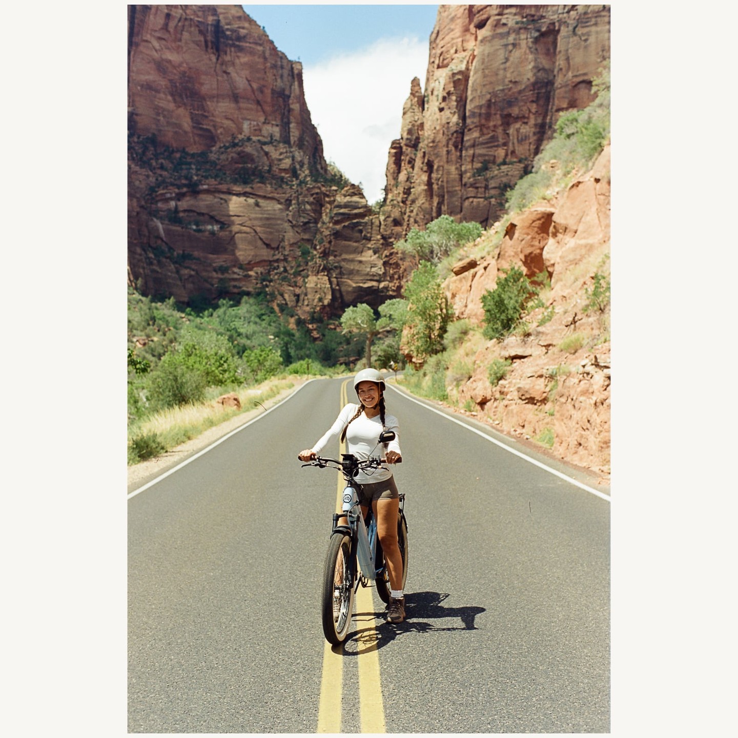 A woman on a bicycle smiles at the camera on a road through a canyon.