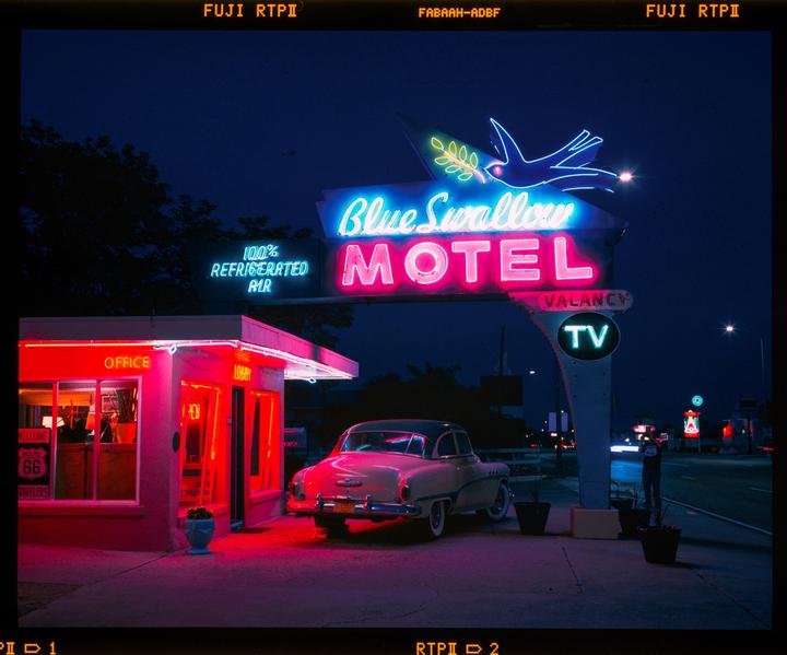 A vintage car is parked in front of the Blue Swallow Motel, illuminated by neon lights at night.