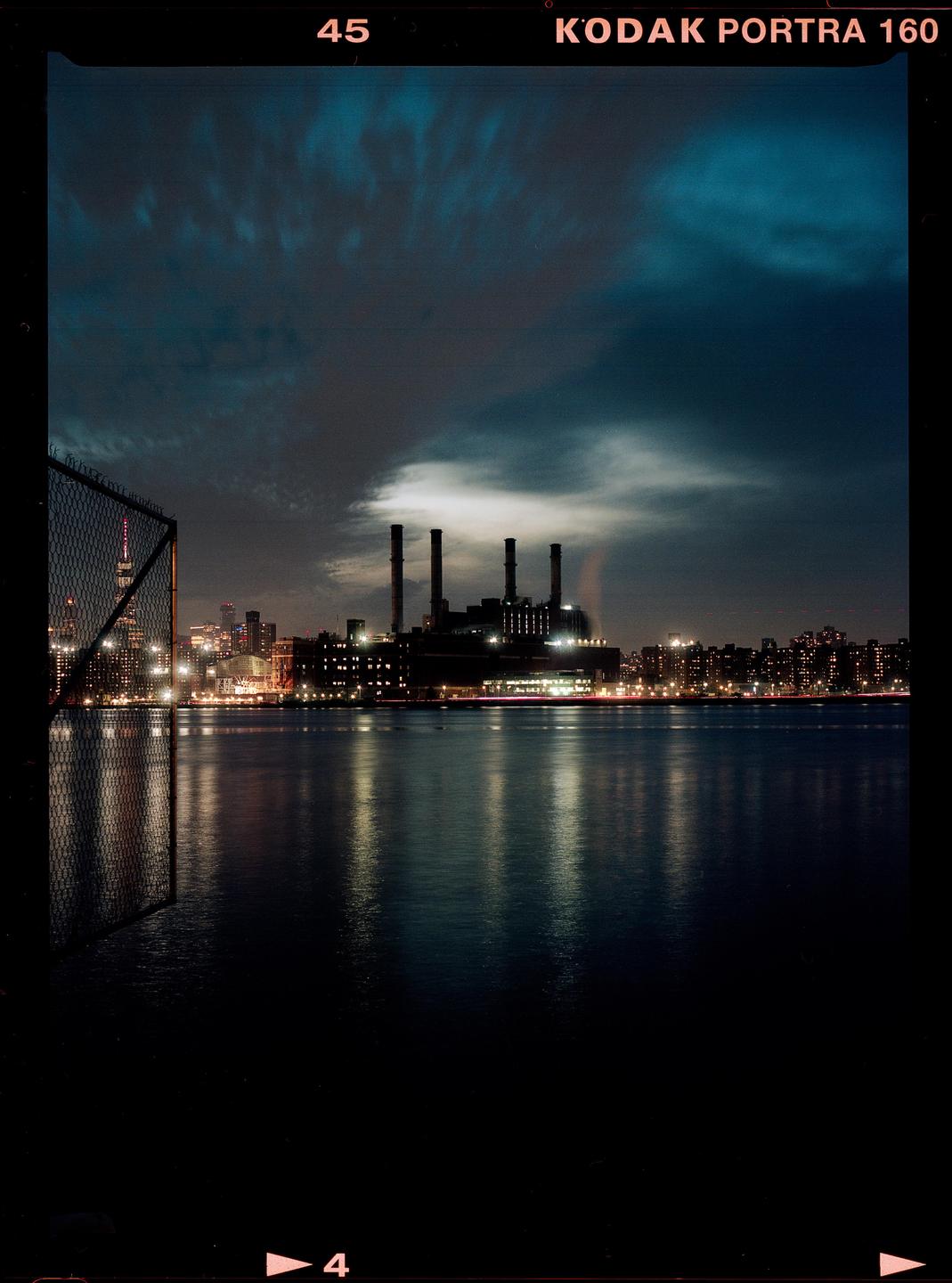 A city skyline with a power plant and its reflection on the water at night.